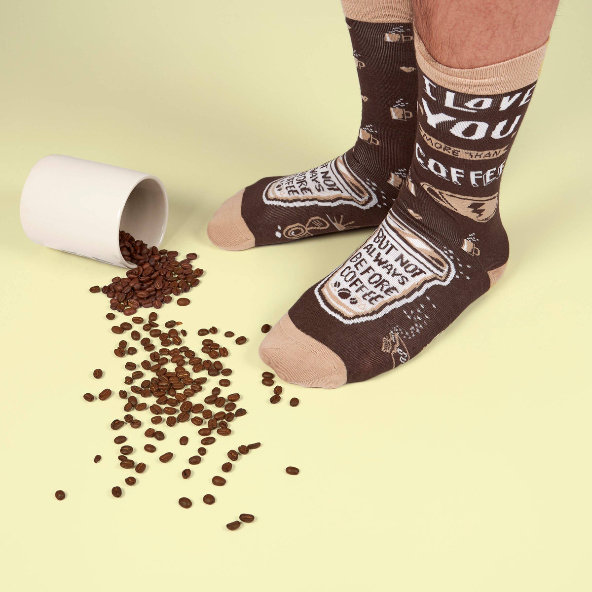 Person wearing socks with coffee-themed text next to a tipped-over cup of coffee beans on a light yellow background