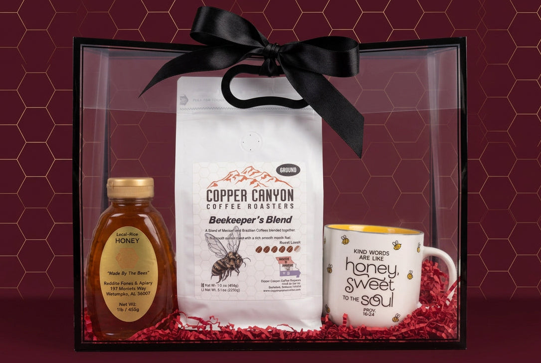 A gift set in a clear plastic box with a black frame and black ribbon, containing a bottle of honey, a bag of Copper Canyon Coffee Roasters 'Bookkeeper's Blend', and a white ceramic mug with bee illustrations and the text 'honey is sweet to the soul'.